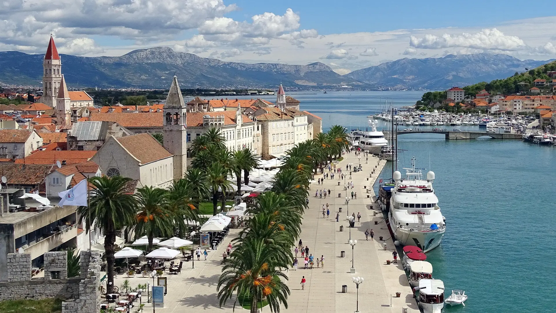 panoramic view of a beachfront town with palm trees, people walking on a promenade and a boat