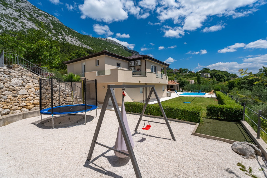 Fenced playground with a trampoline, a swing, badminton plot, and grass mini golf, a lounge sitting area