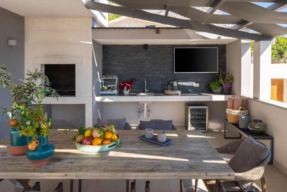 An outdoor dining area with a dining table for 8 people with a barbecue, a TV and a fridge