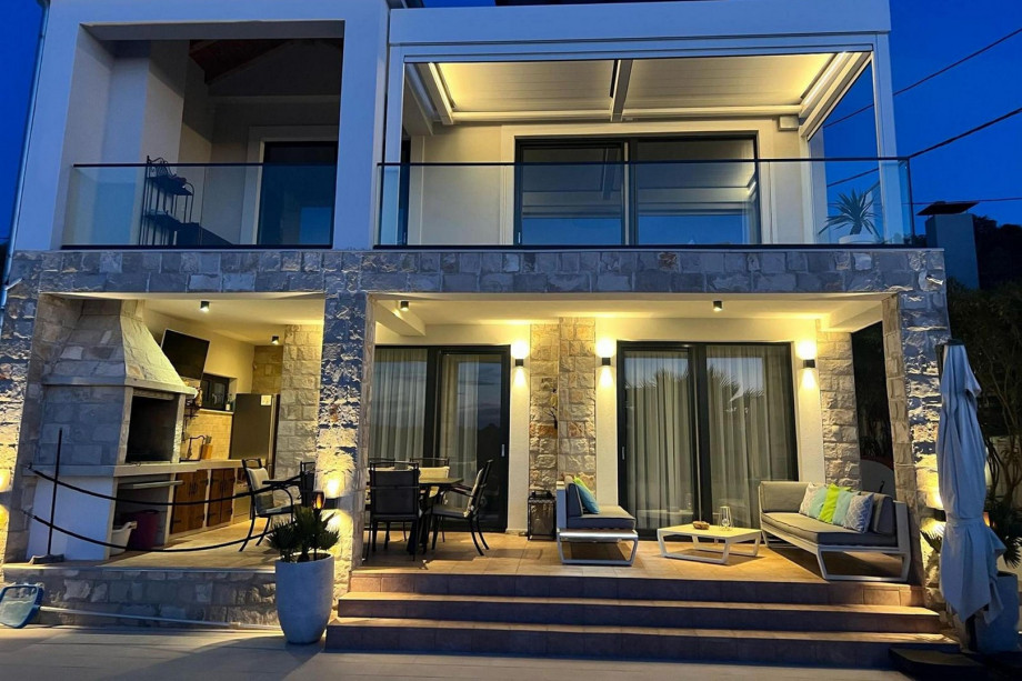 A beautifully lit modern villa with a newly covered terrace, stone accents, and a cozy outdoor seating area with a barbecue creates an inviting ambiance at dusk.