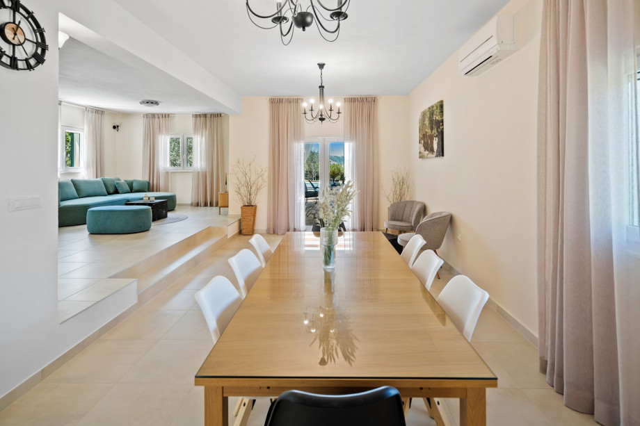 A bright, elegant open-plan dining and living area features a large wooden table, modern white chairs, and a cozy lounge with a green sectional sofa.
