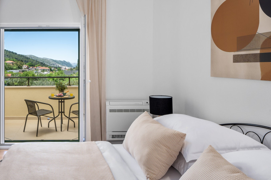 A stylish bedroom with a cozy bed and soft neutral tones, opening onto a balcony that offers picturesque views of green hills