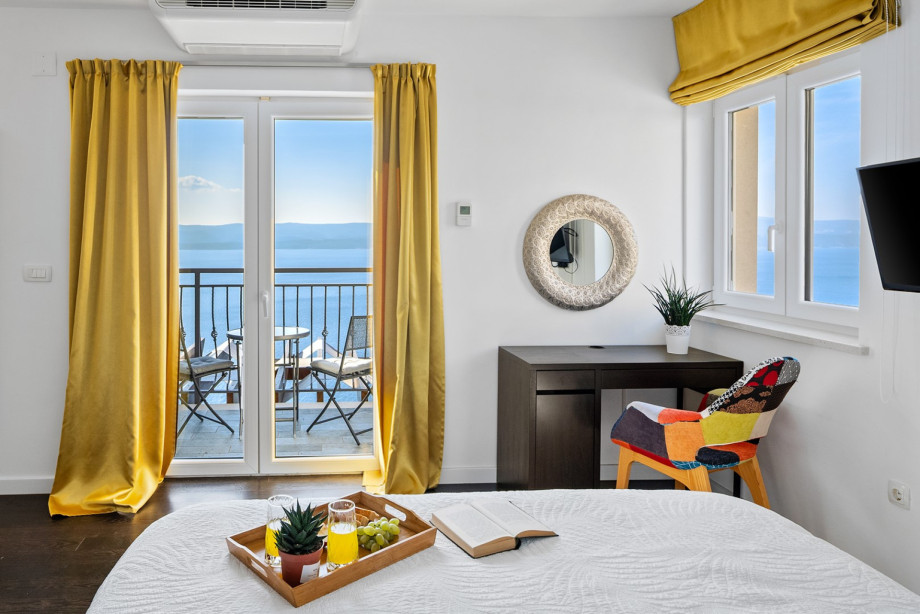 This yellow-accented bedroom features balcony access with sea views, a comfortable bed, and a mirrored desk area