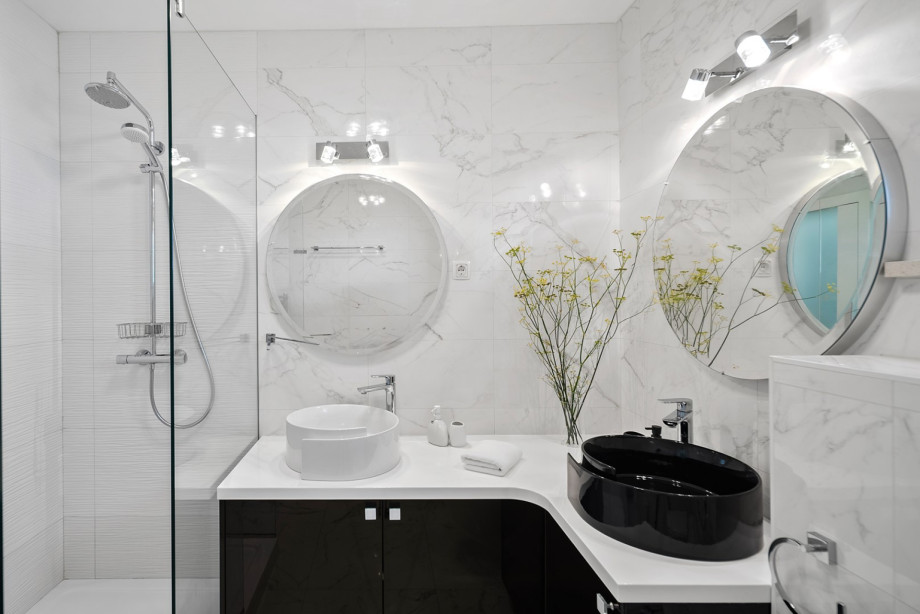 Elegant, marble-finished bathroom highlights the dual vessel sinks and illuminated round mirrors, separated by a glass shower wall