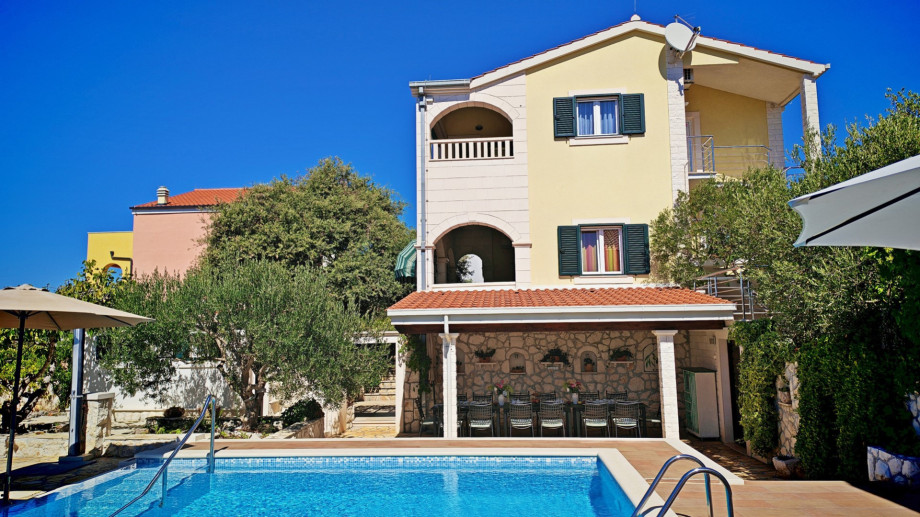 The villa's main building, featuring three separate apartments and an arched balcony, overlooks the pool area and the shaded, stone-clad outdoor dining space.