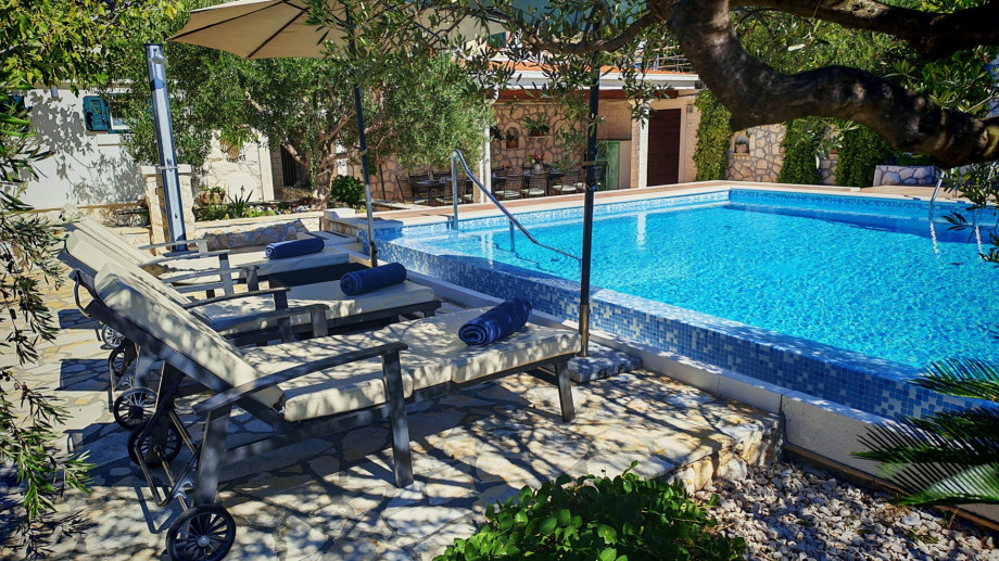Comfortable lounge chairs are perfectly positioned on the sun deck under the shade of olive trees, ready for relaxation beside the sparkling blue pool.