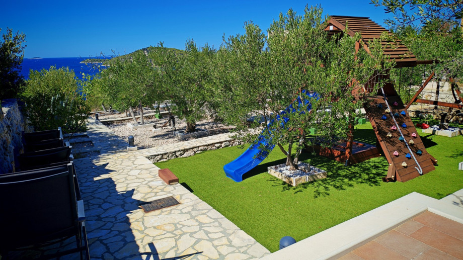 The fun zone for kids, nestled among olive trees with a slide and climbing frame, provides entertainment with a beautiful sea backdrop.