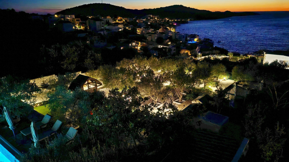 An elevated view at sunset shows the villa's illuminated grounds and olive trees contrasting beautifully with the sparkling coastline.