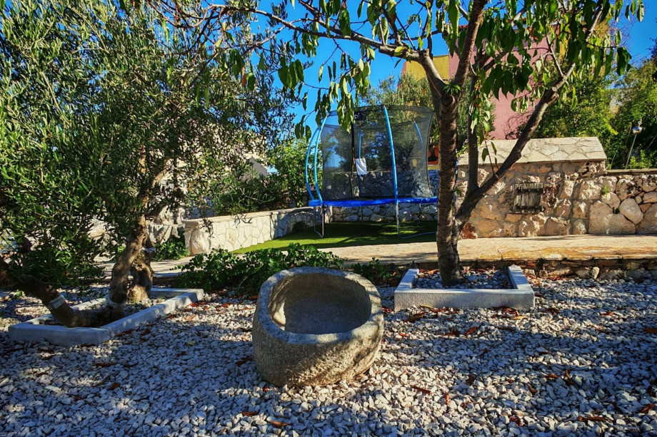The dedicated kids' fun zone features a trampoline surrounded by olive trees and a pebble path, ensuring hours of active outdoor entertainment.