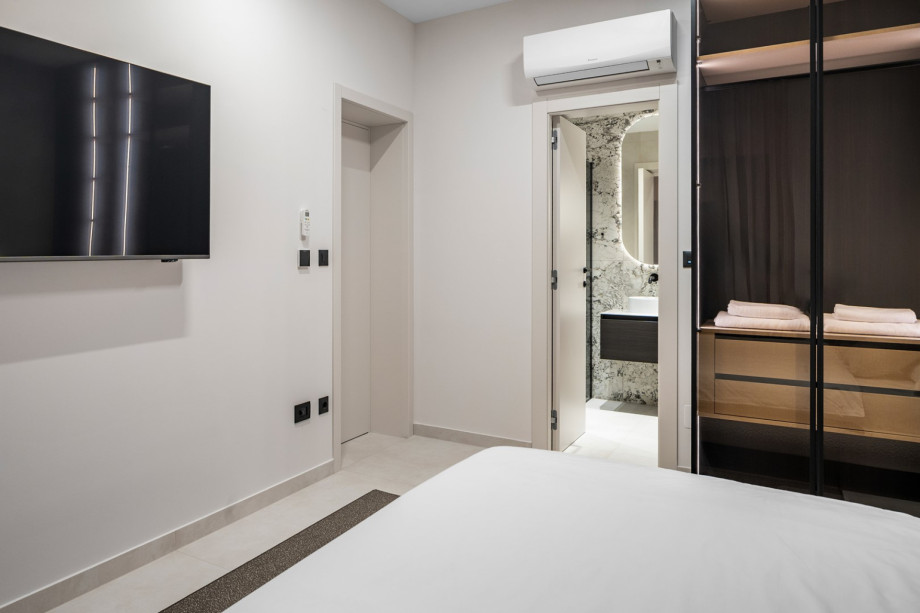 A view of a bedroom featuring a wall-mounted TV, air conditioning, and a doorway leading to an en-suite bathroom with marble accents.