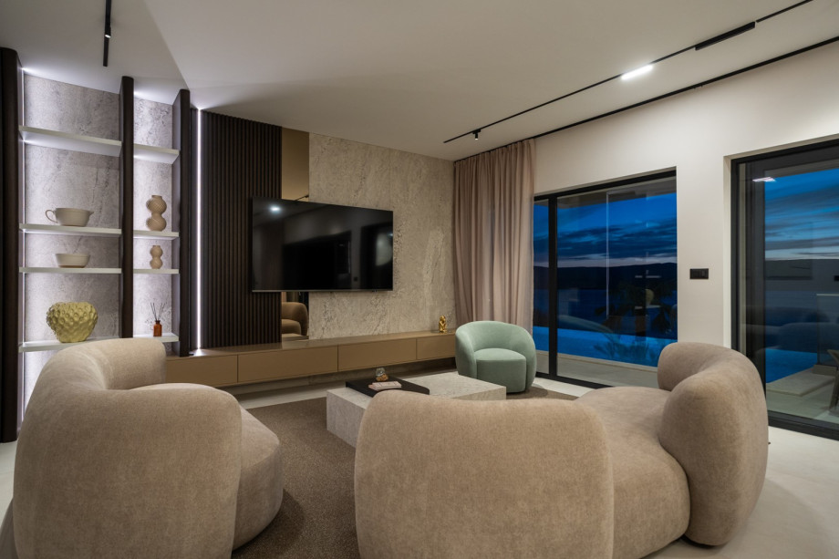 The indoor living room, spanning 65 sqm at pool level, features comfortable sofas and a TV, connecting seamlessly to the outside via large glass doors.