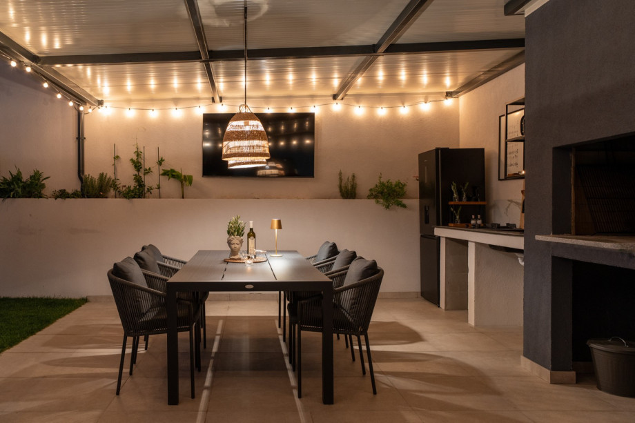 Covered summer kitchen with al fresco dining for 6, a TV, and a traditional barbecue area, illuminated at night.