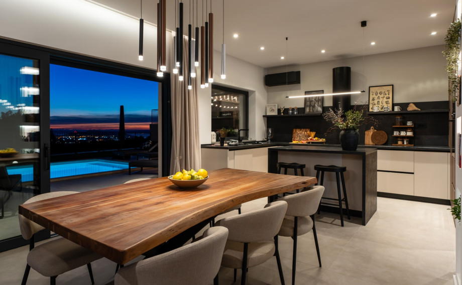 Open-plan dining and kitchen at twilight, with light reflecting off the pool and the distant city lights.