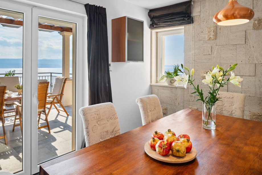 The indoor dining area, featuring stone-effect walls and modern furnishings, connects directly to the outdoor balcony with dining furniture and sea views