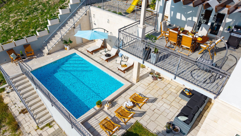 A wide aerial view shows the entire outdoor area, including the pool, sun deck, children's playground, and the shaded dining terrace