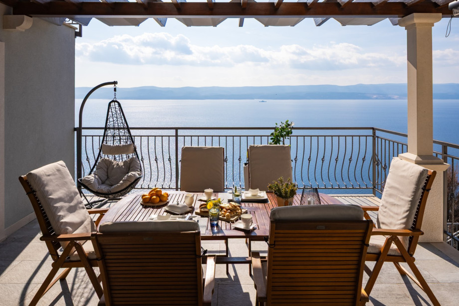 The outdoor dining terrace features a large wooden table and a hanging rattan chair, all set against a stunning backdrop of the open sea and islands