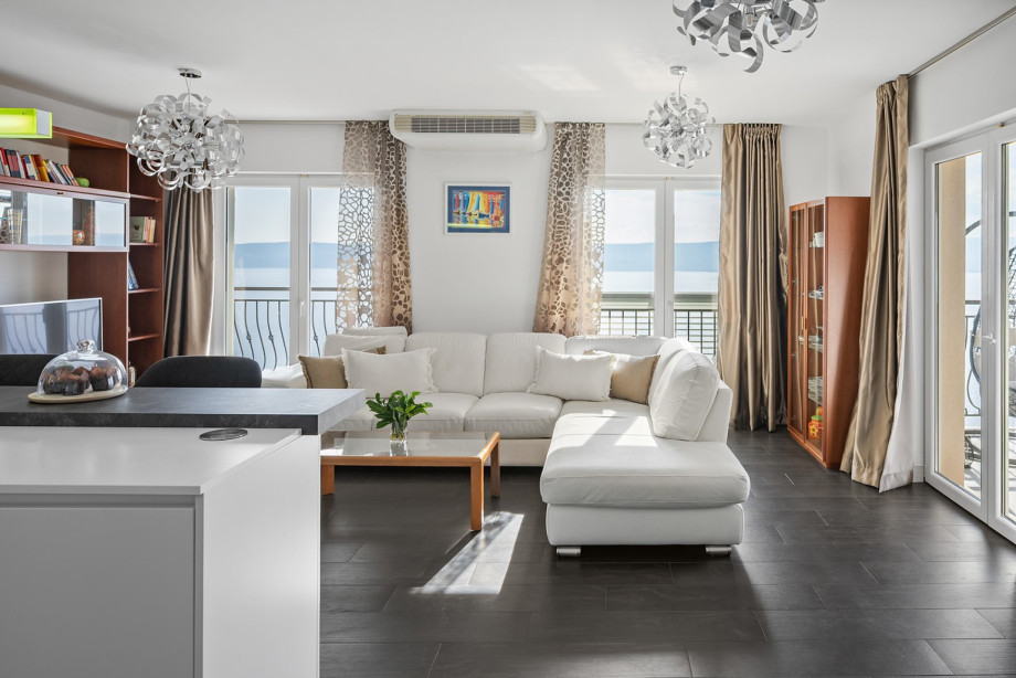 The bright, air-conditioned living room is furnished with a white sectional sofa, modern chandeliers, and large glass doors that offer direct sea views and balcony access
