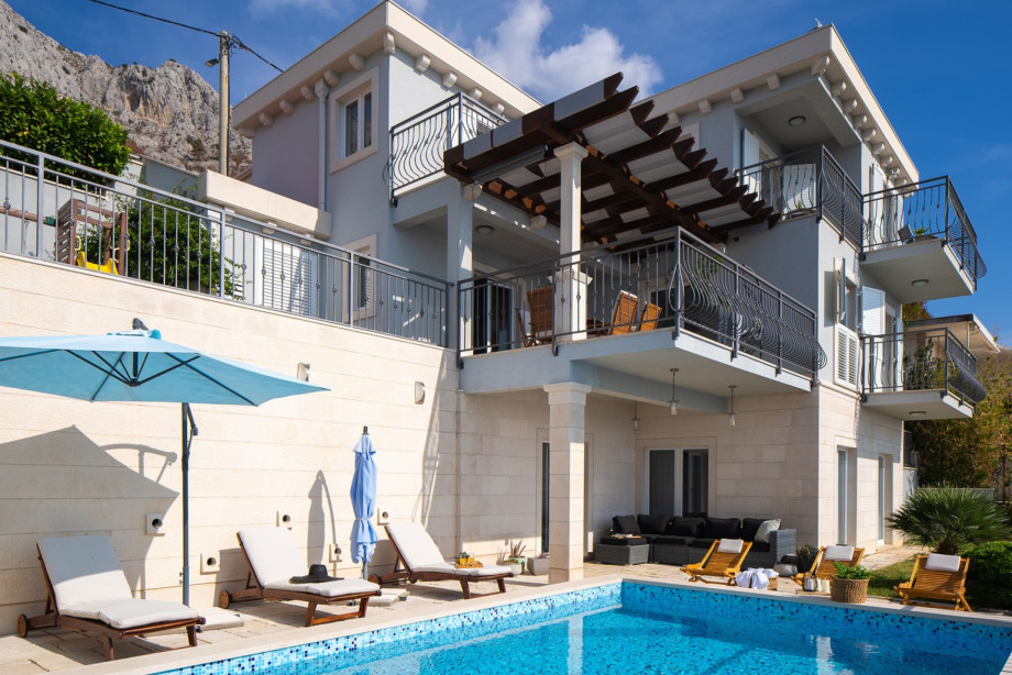 A full view of the villa from the pool level shows the modern architecture, shaded terraces, and the prominent mountain ridge above