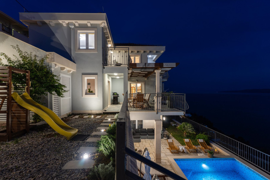The villa exterior is beautifully lit at night, highlighting the architecture and the private playground with a yellow slide and ambient lighting