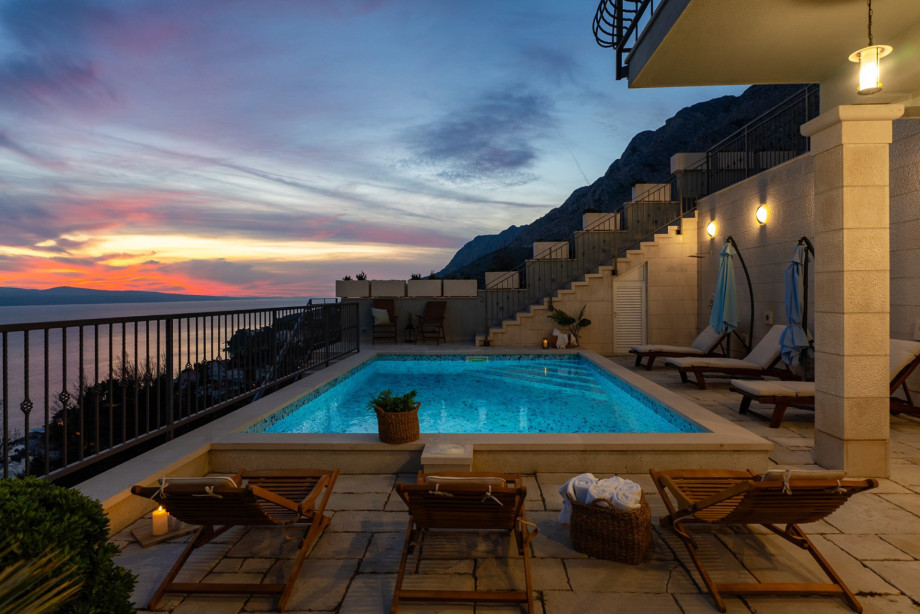 Wooden sun loungers and comfortable seating invite guests to relax by the glowing pool as the stunning colors of the sunset fade into the night