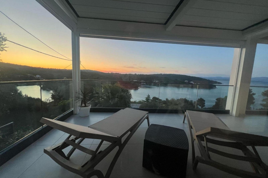 A tranquil view from a newly-roofed terrace, featuring lounge chairs and glass railings, overlooking a peaceful bay and lush greenery as the sun sets in the distance.