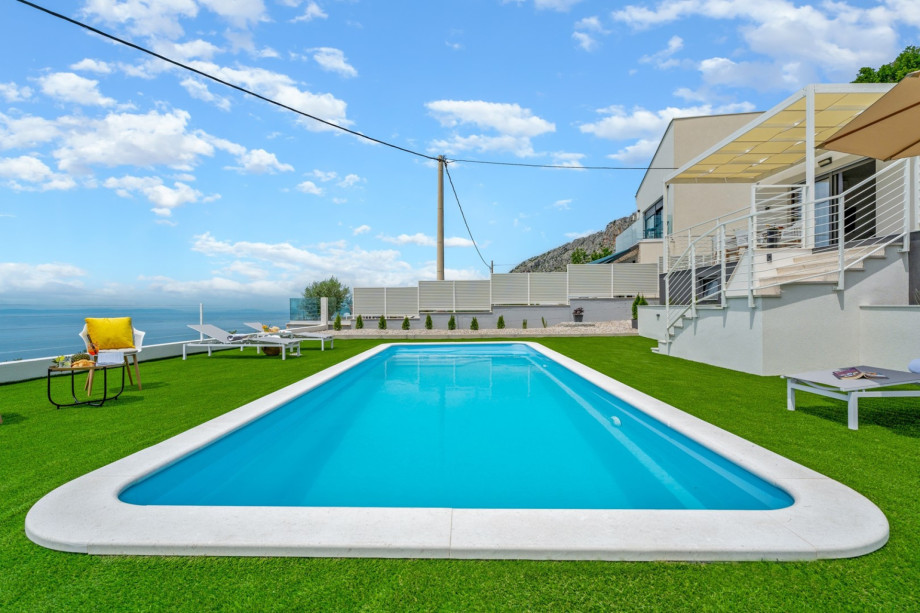 A sun deck area with artificial grass and with 4 deck chairs, a lounge corner, an outdoor shower, and a sun umbrella