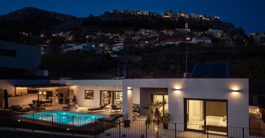 Villa exterior at night, with the illuminated pool and the historic Klis Fortress in the background.