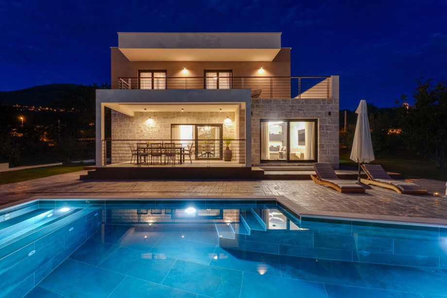 The illuminated villa exterior showcases the spacious stone terrace and crystal-clear swimming pool.