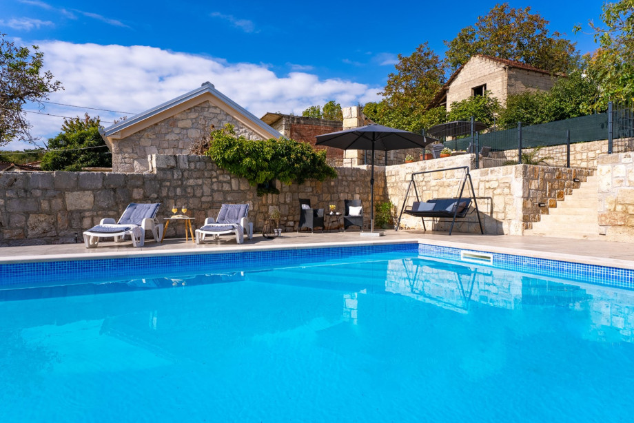 The pool's edge provides a refreshing view of the tiered sun deck, featuring loungers, an umbrella, and a swing chair, alongside the rustic stone structure.
