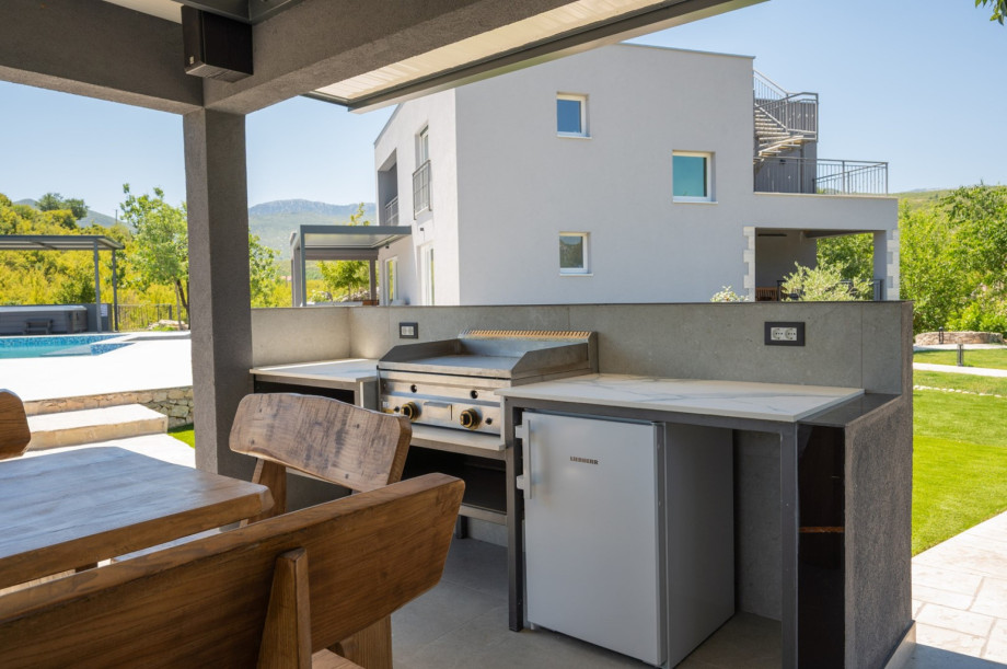 Outdoor dining area featuring a gas BBQ, fridge, and sink next to the lawn and pool area.