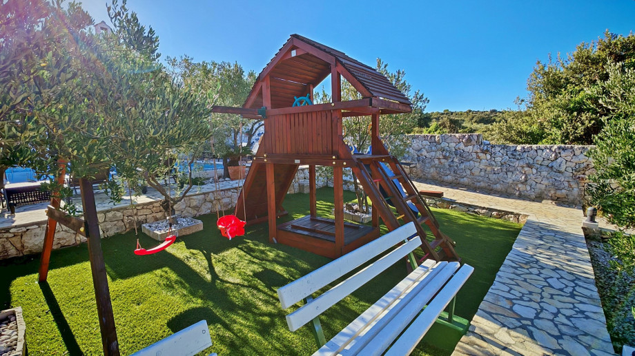 Another view of the kids' play area, showcasing the wooden climbing frame, swings, and a bench, surrounded by stone walls and greenery.