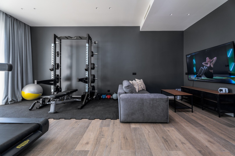 Fitnessstudio mit Klimaanlage und Ausgang zum Poolbereich, verbunden mit einem Spielbereich mit Sofa.