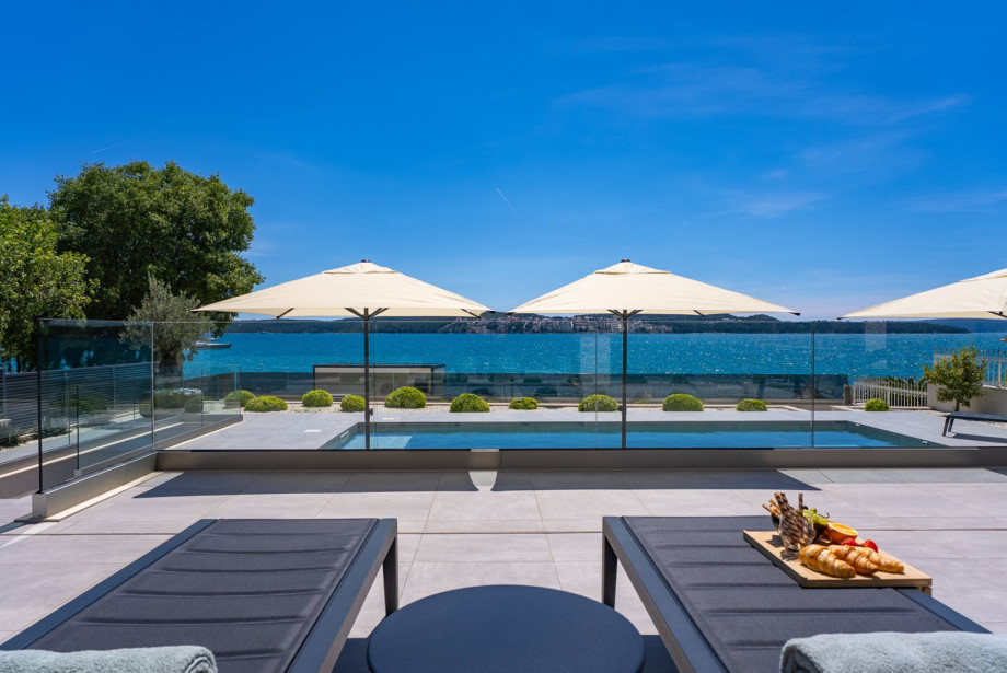 A modern seaside terrace with comfortable sun loungers, elegant umbrellas, and a view of the crystal-clear blue sea.