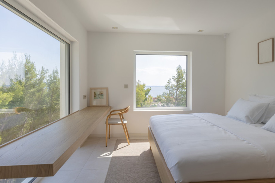 Helles Schlafzimmer mit einem bequemen Bett, einer langen Arbeitsfläche aus Holz und großen Fenstern mit Blick ins Grüne und auf das Meer