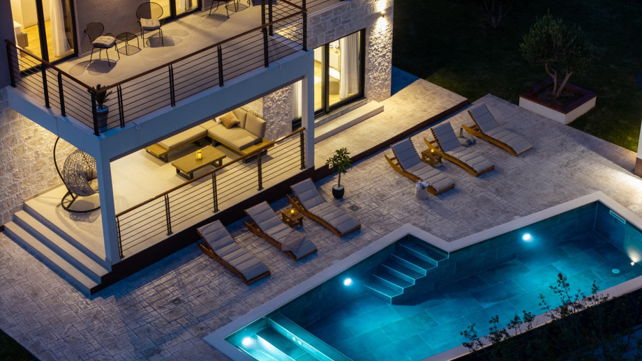 The illuminated villa exterior showcases the spacious stone terrace and private swimming pool.