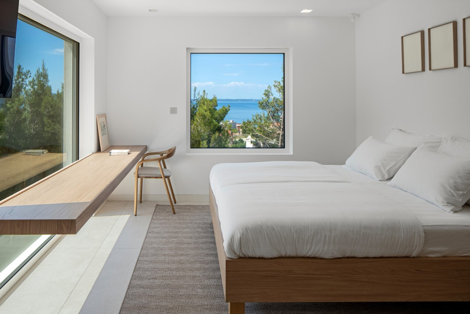 Bright bedroom with a comfortable bed, long wooden workspace, and large windows framing views of greenery and the sea