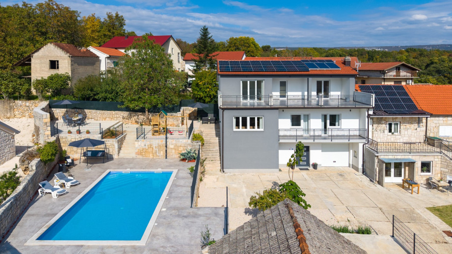 The modern villa exterior, featuring balconies and solar panels, overlooks the 50sqm private swimming pool and the tiered sun deck and playground area.